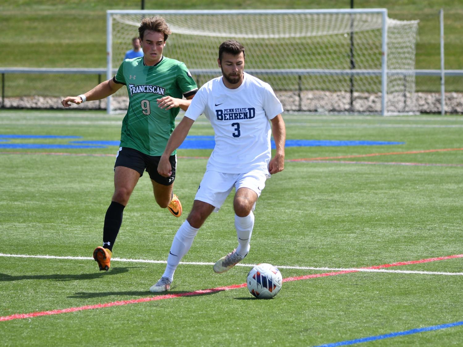 A Penn State Behrend men's soccer player advances the ball to midfield while being pressured by a defender.