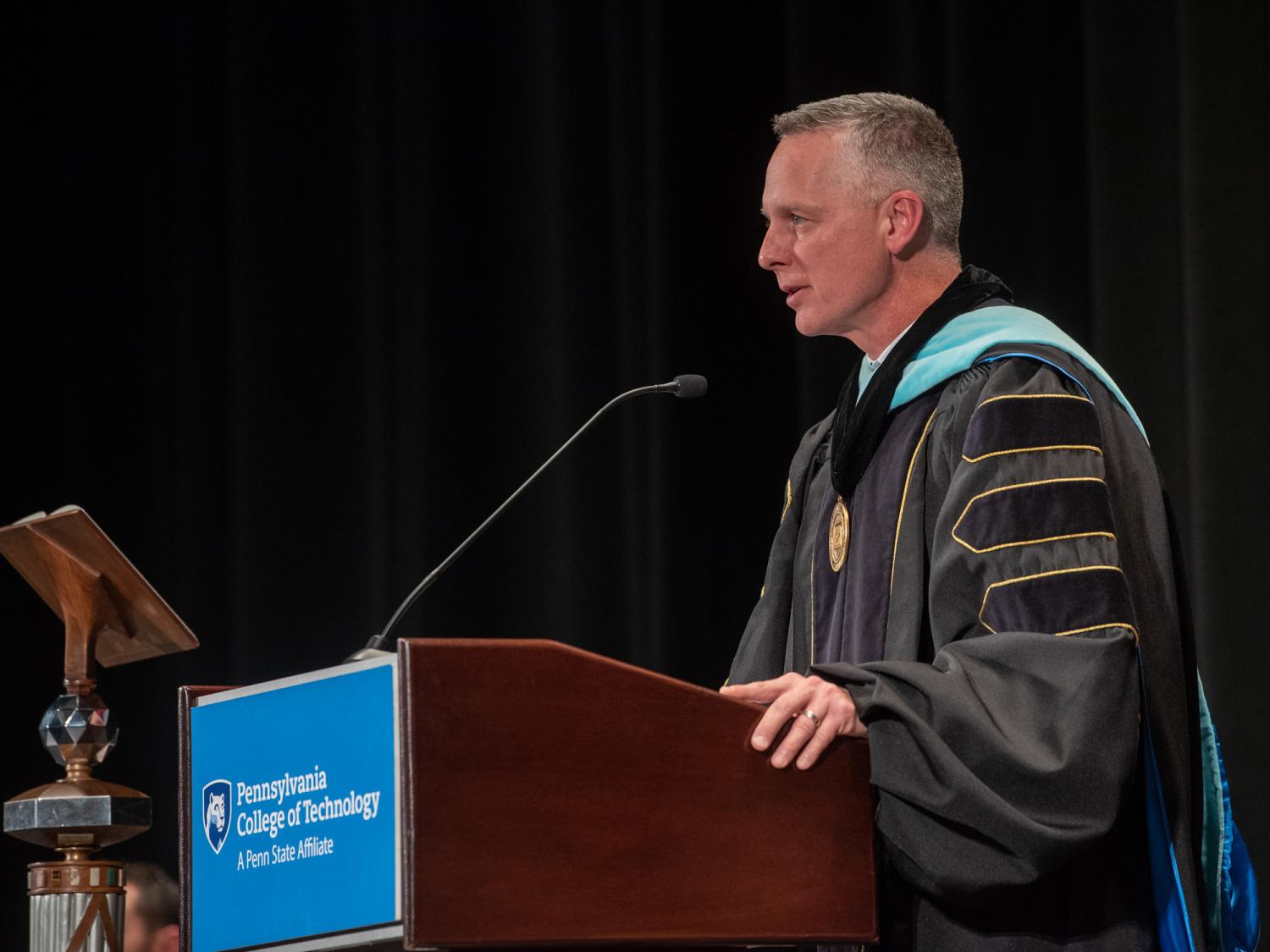 Michael Reed speaking at a podium with a Penn College logo and a dark background