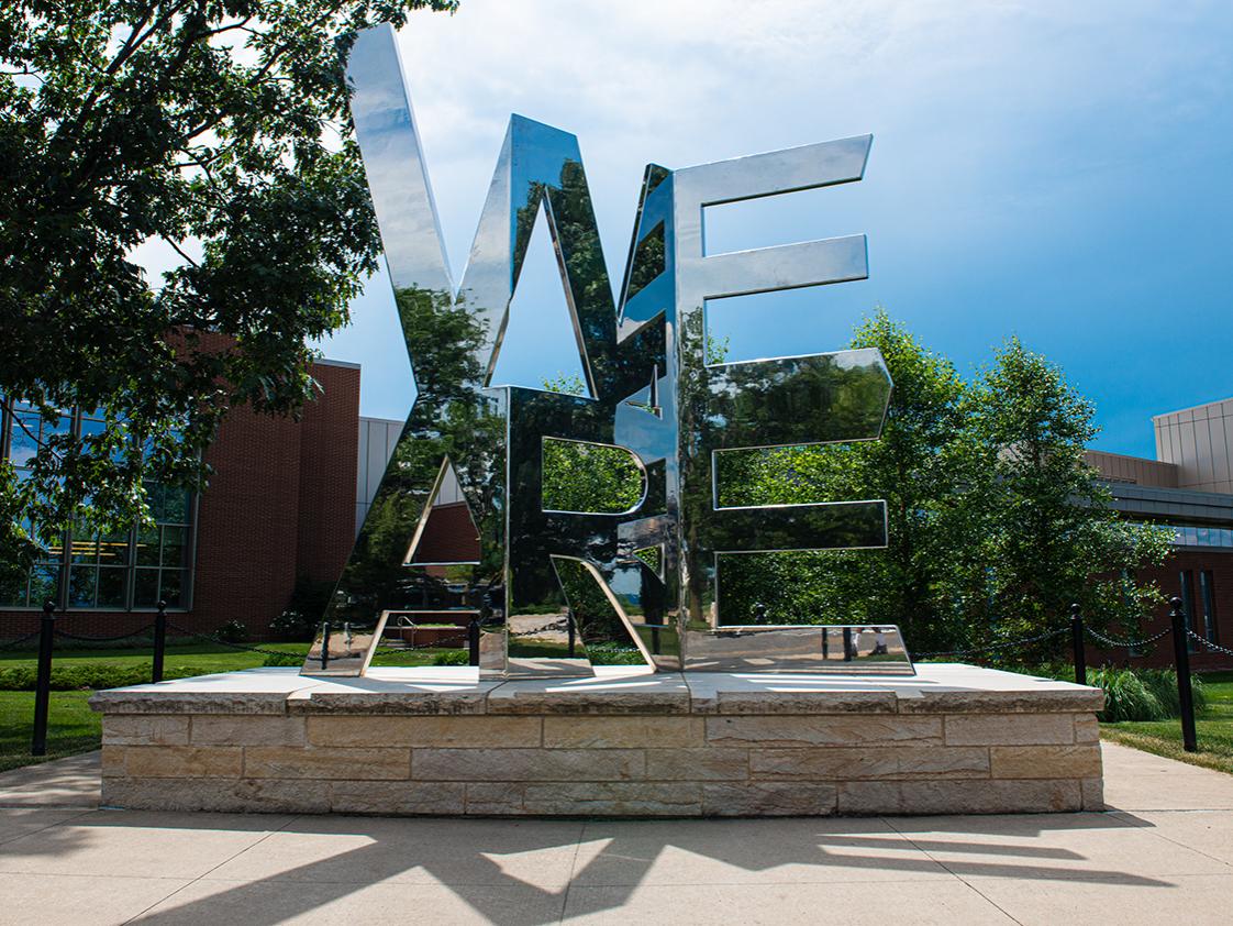 mirrored "we are" word sculpture against backdrop of brick building and blue sky
