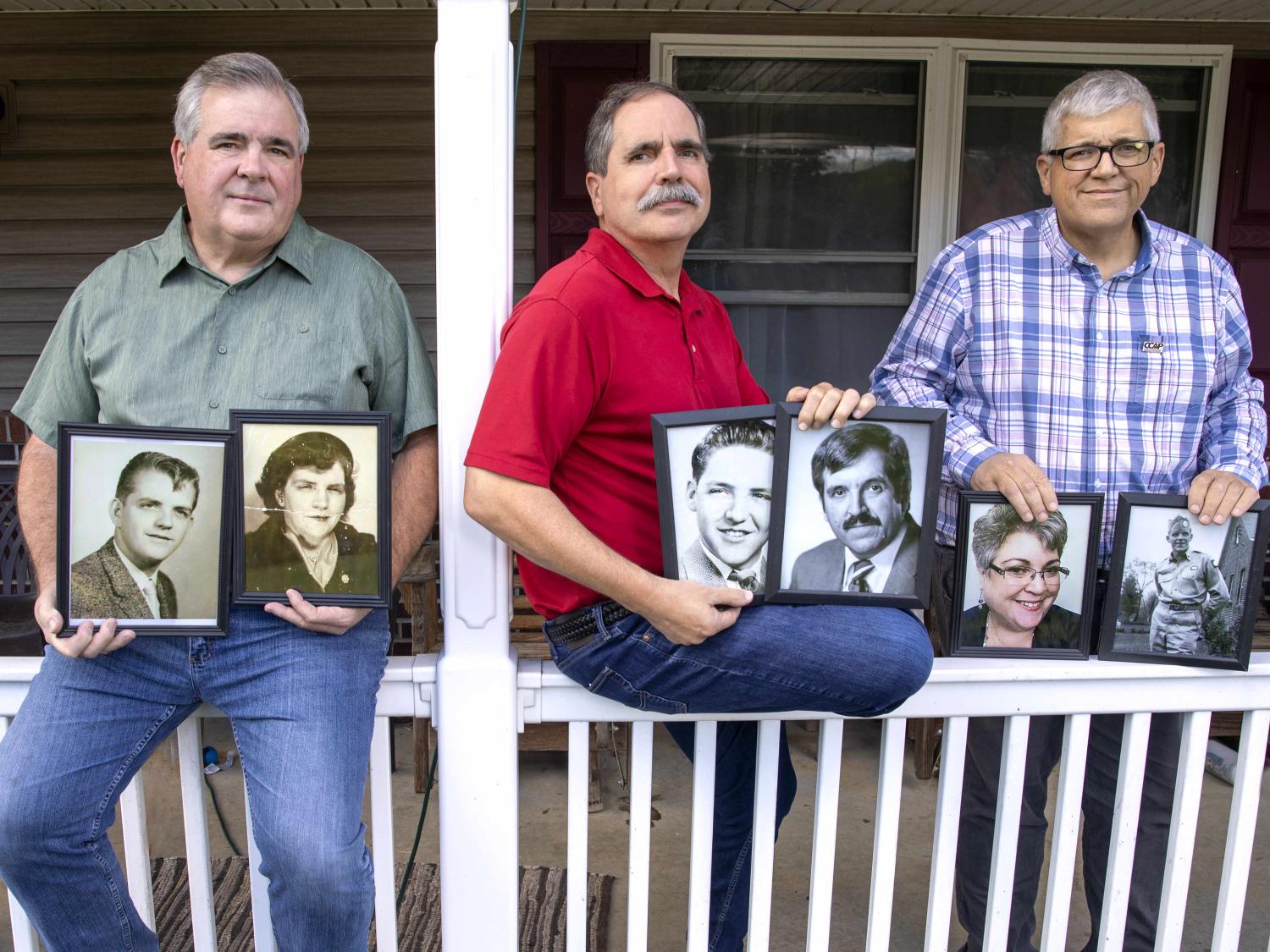 Three men sit and lean on a porch railing, holding framed, black-and-white photos.