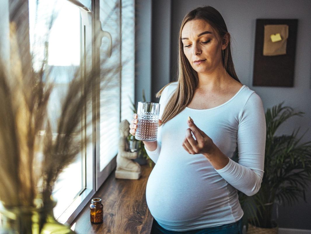 A pregnant woman stands by the window with a pill in her left hand and a drink in her right.