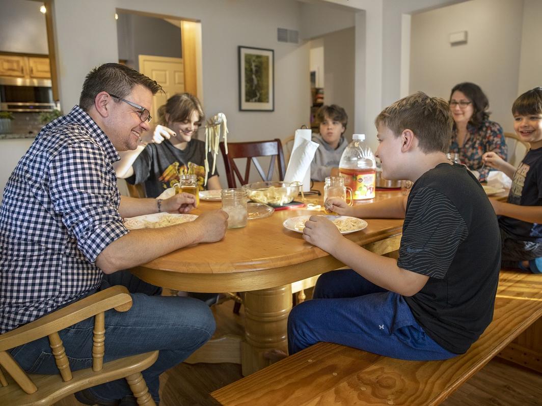 A man, woman and four children sit around a table eating a meal. Many of them are smiling.