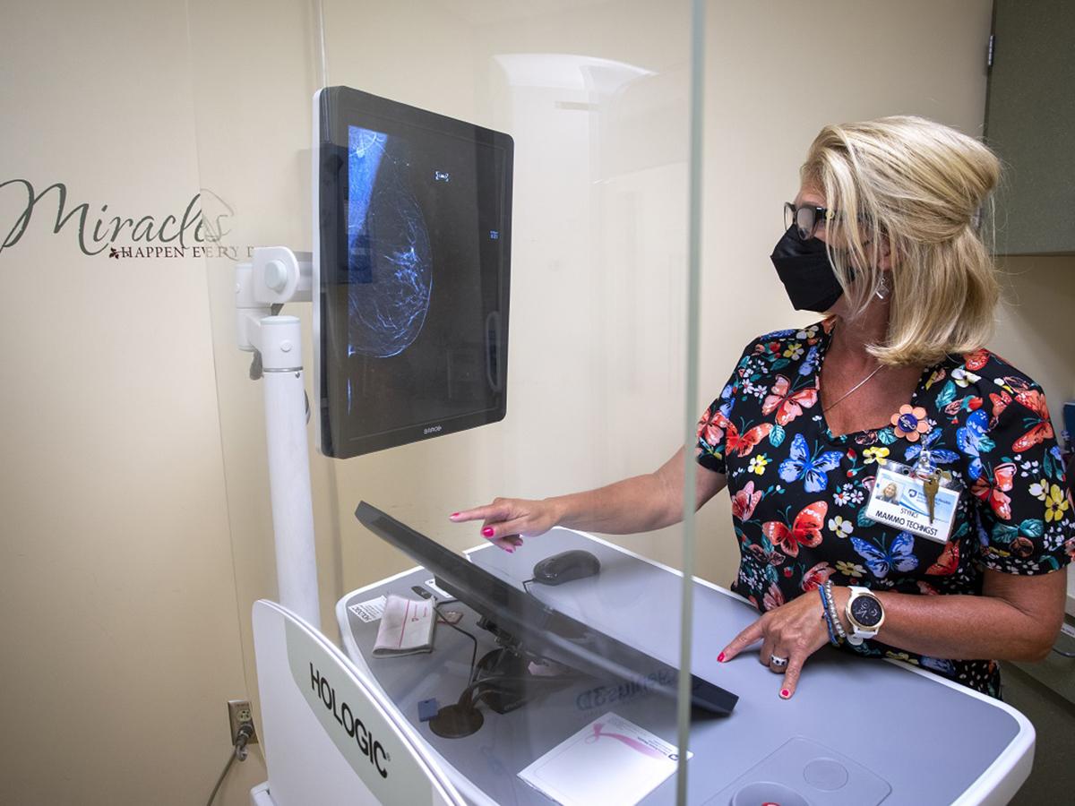 A woman works at a computer terminal in a medical exam room. On the wall next to her are the words “Miracles happen every day.” On the screen is a X-ray image of a breast.