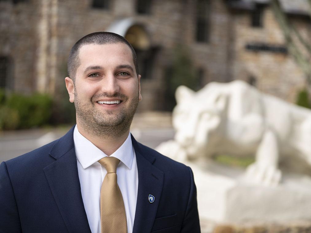 Man in a suit and tie smiling in front of Nittany Lion statue.