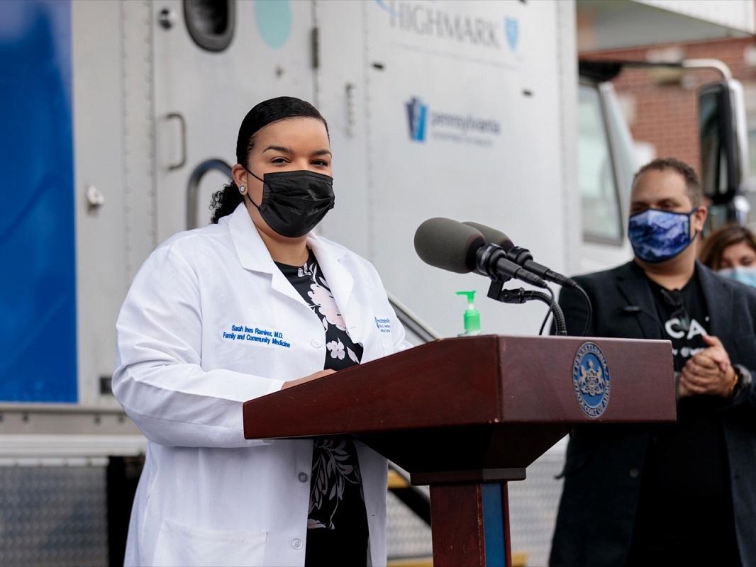 Dr. Sarah Ramirez of Penn State Health stands at a podium next to a man and woman during a press conference at Beacon Clinic in Harrisburg. She is wearing a white coat and a face mask. Behind her is a van with the Highmark logo on it.