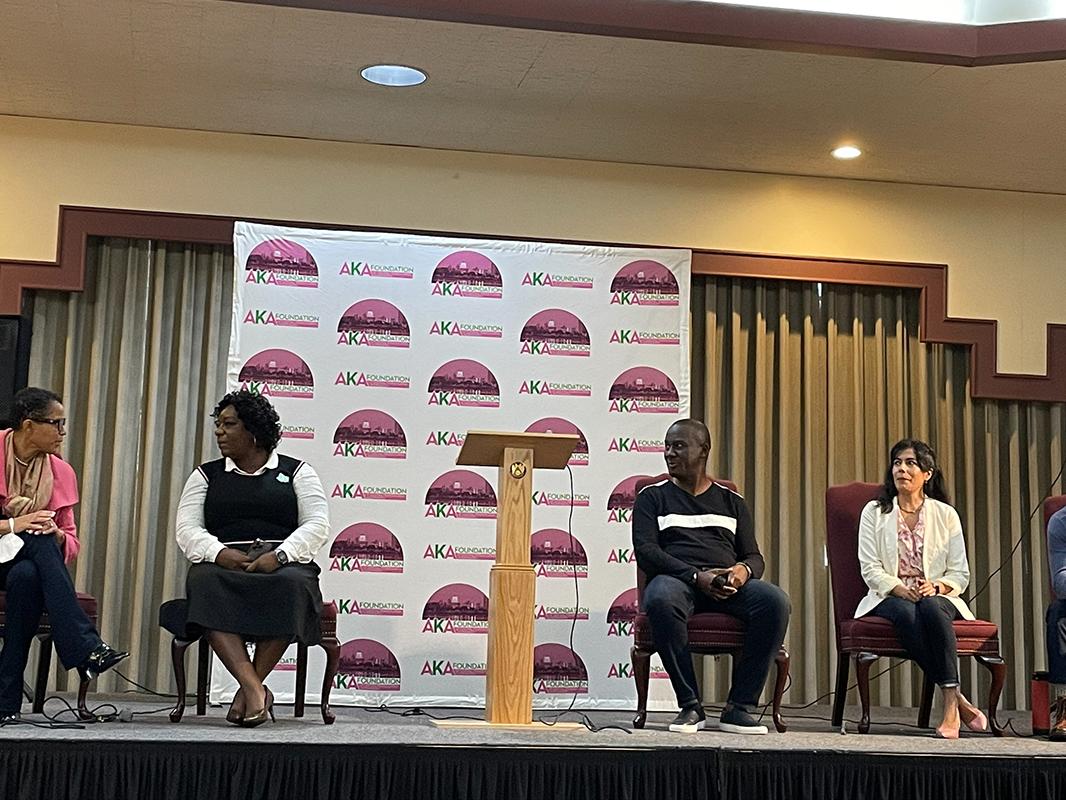 A row of five people sit on a stage. At the center is a podium and behind them a logo appears on a poster.