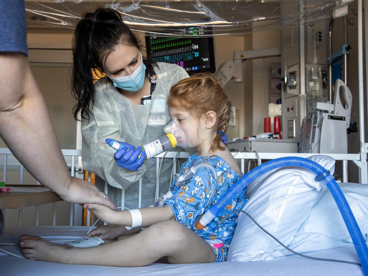A health care worker in a mask, gown and gloves holds a breathing apparatus to the mouth and nose of a little girl, who is sitting up in a hospital bed.
