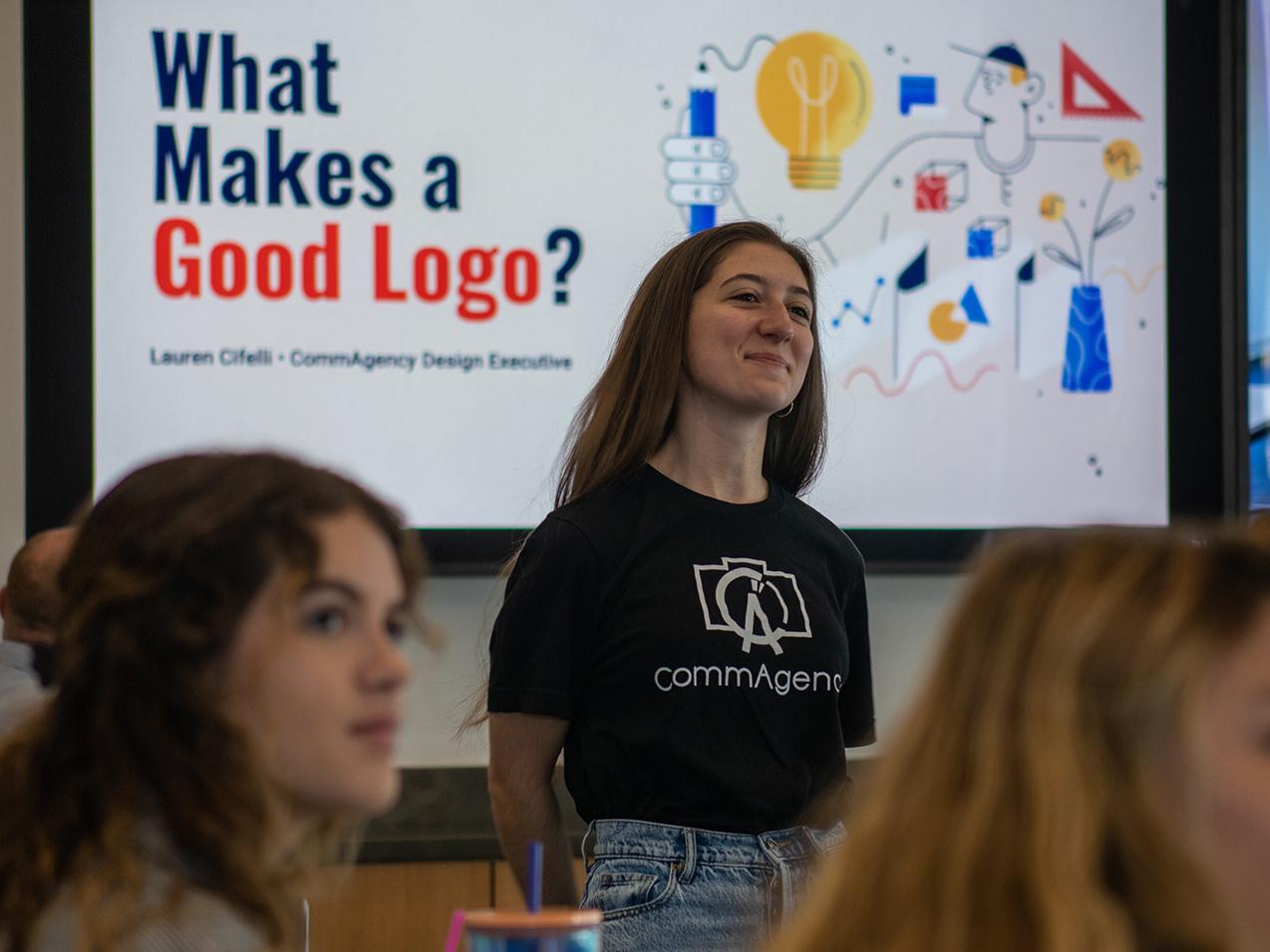 A student stands to speak with a screen on the back showing the words "What Makes a Good Logo?"