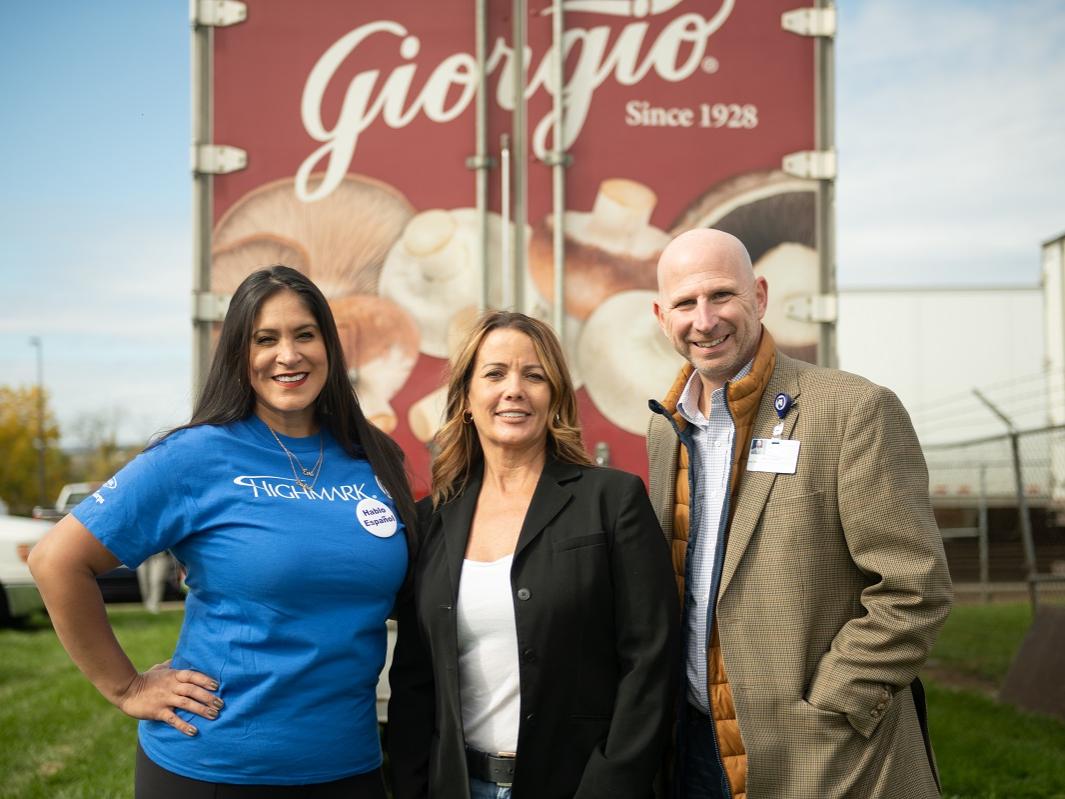 Two women and a man stand next to each other in front of a Giorgio truck. There are pictures of mushrooms on the truck. In the background on the left, cars are parked in the grass. On the right, trucks can be seen behind a fence.
