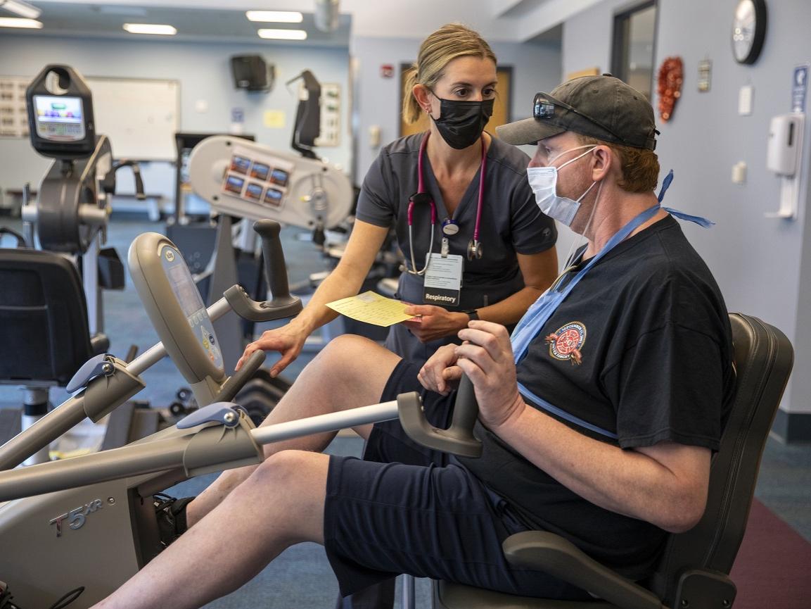 A man in a ball cap and shorts works on a piece of equipment in a gym. A woman in a surgical mask stands next to him.