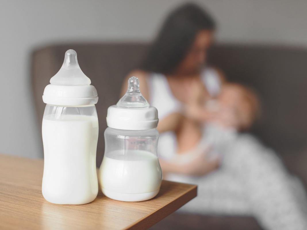 Two bottles of breast milk sit on a counter. Faded into the background, a mother breastfeeds her baby. 