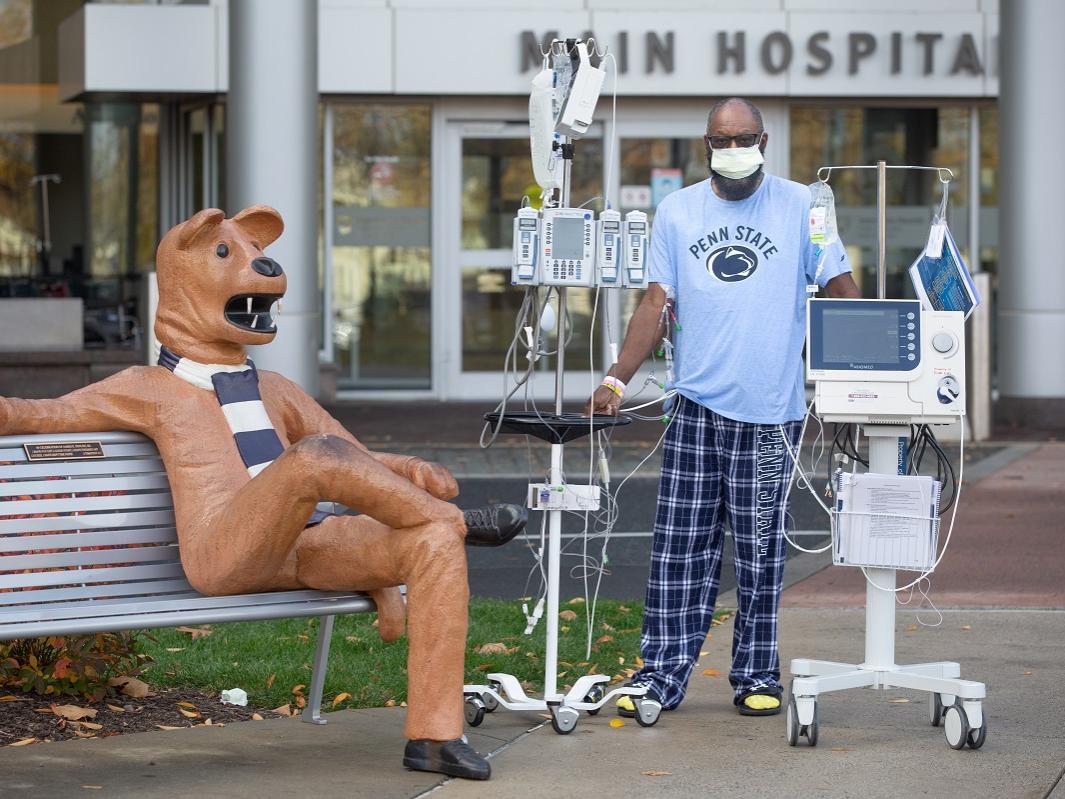 Eddie Holman stands in front of Penn State Health Milton S. Hershey Medical Center with an IV pole and monitor. He is wearing a Penn State T-shirt and pants, a surgical mask and glasses. Next to him is a statue of the Penn State Nittany Lion sitting on a bench.