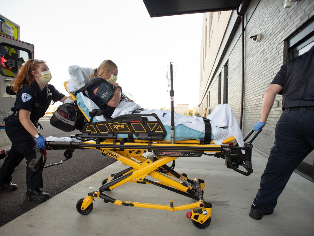 A woman sits upright on a litter as two ambulance staff members guide it from the ambulance to the sliding door entryway of a hospital.