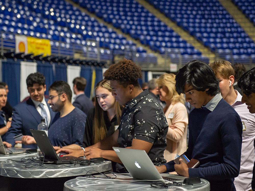 People stand at laptop computers. stadium seating is visible in the background. 