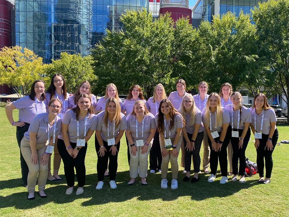 Team of 18 young women wearing light purple polo shirts and lanyards pose with city buildings in background.