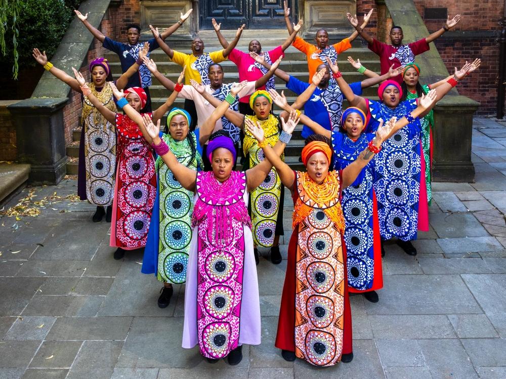 Choir members dressed in boldly patterned gowns and head scarves stand in a diamond shape as each holds their arms up in a V formation.