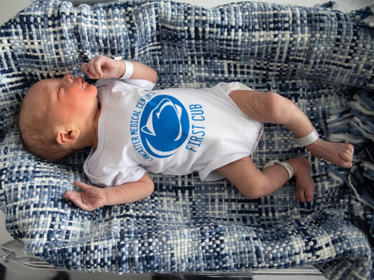 A newborn baby lays on a textured blanket. He wears a onesie with a Penn State logo and the words “Lancaster Medical Center’s First Cub.”