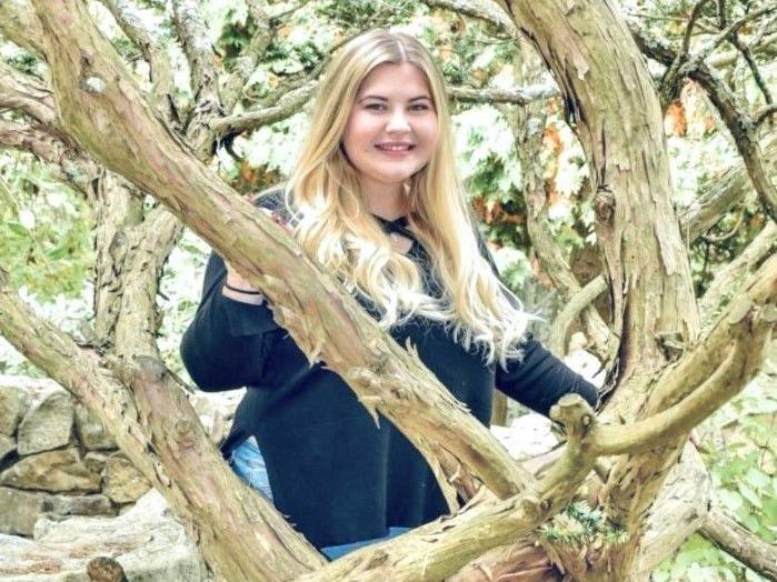 Blonde woman in a dark sweatshirt posing from behind tree branches.