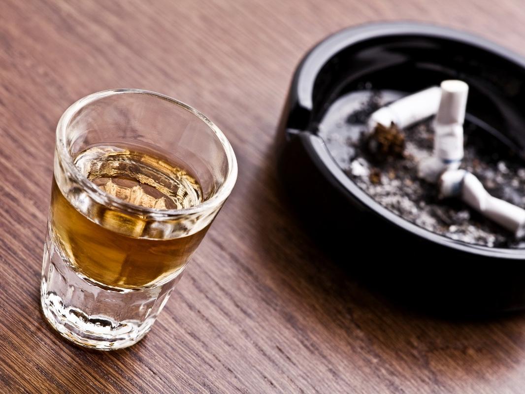 A shot glass of whiskey sits next to an ashtray with three discarded cigarettes. 