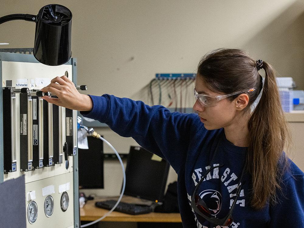 Young dark-haired woman with ponytail and safety glasses adjusts equipment knobs in laboratory.