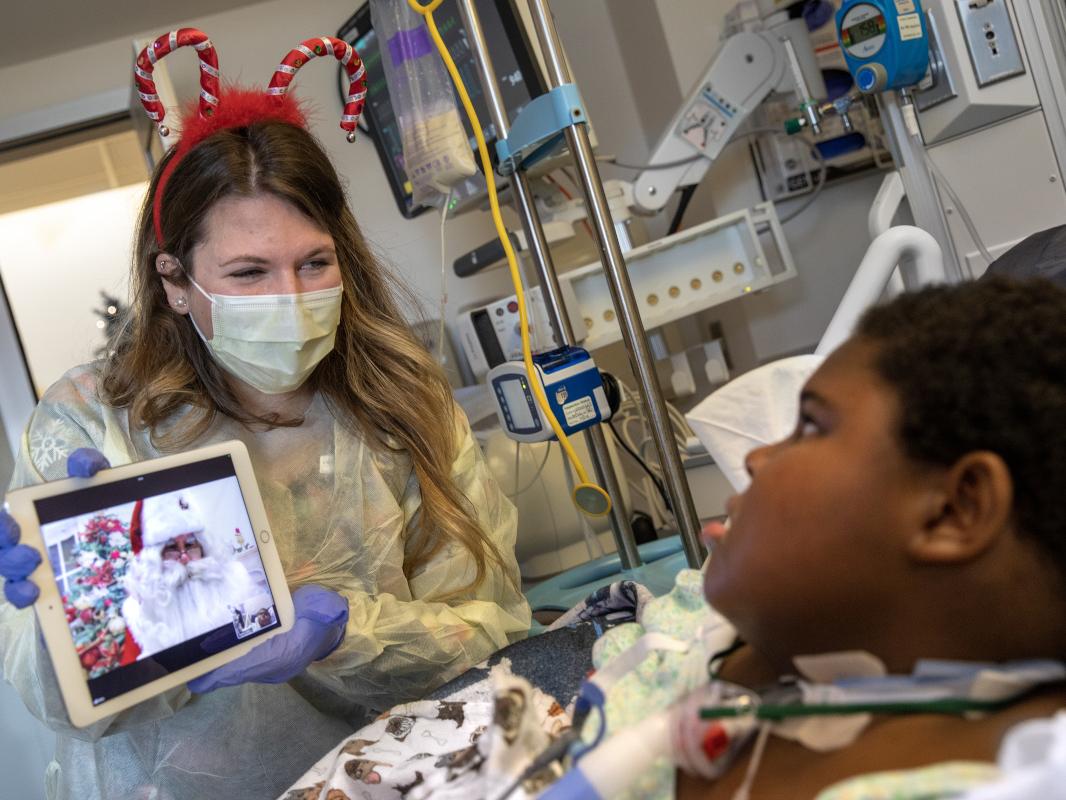 A woman in a candy cane headband, surgical mask and other personal protective equipment holds an iPad that bears a likeness of Santa Claus, while a young male patient looks on from his hospital bed.