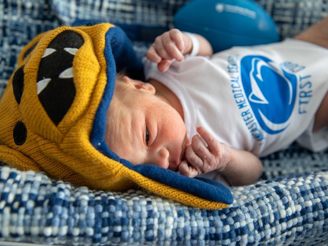 A baby lies on a cushion. He wears a Nittany Lion hat and a onesie with the Penn State logo.