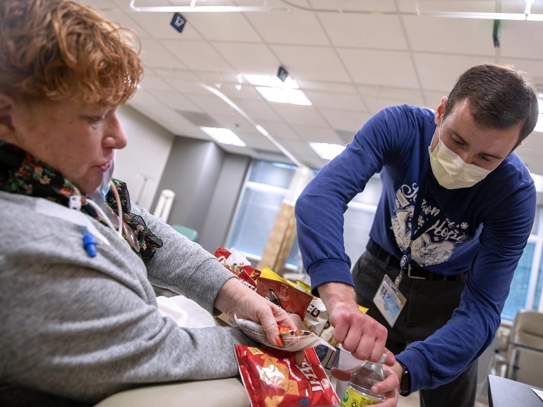 A man in a surgical mask an and a long-sleeve T-shirt opens a bottle for a woman seated in a chair.