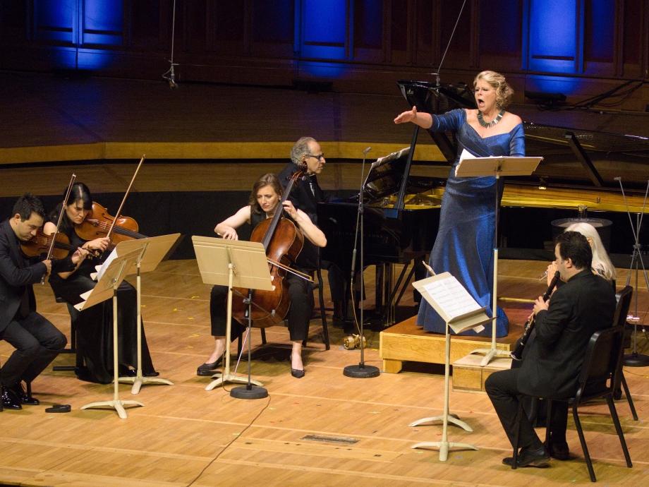 A classical music ensemble sits in a semi-circle while a vocalist wearing a formal gown sings.