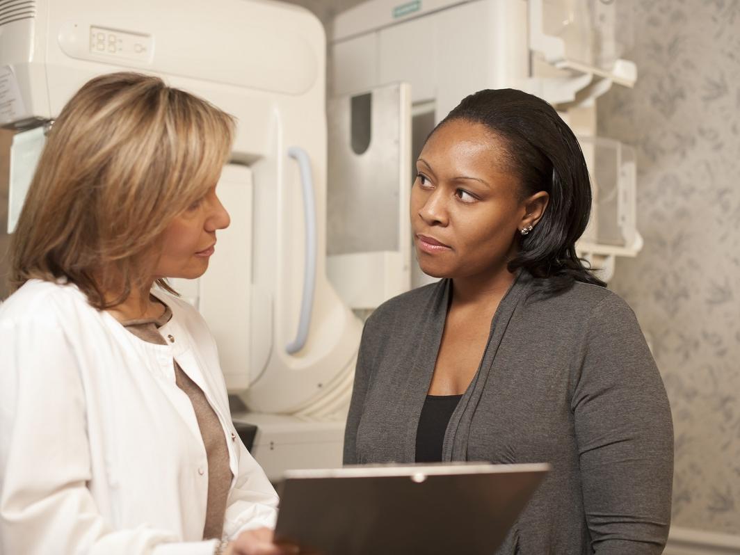 A technician explains a mammogram to an adult woman patient. A mammogram machine is in the background.
