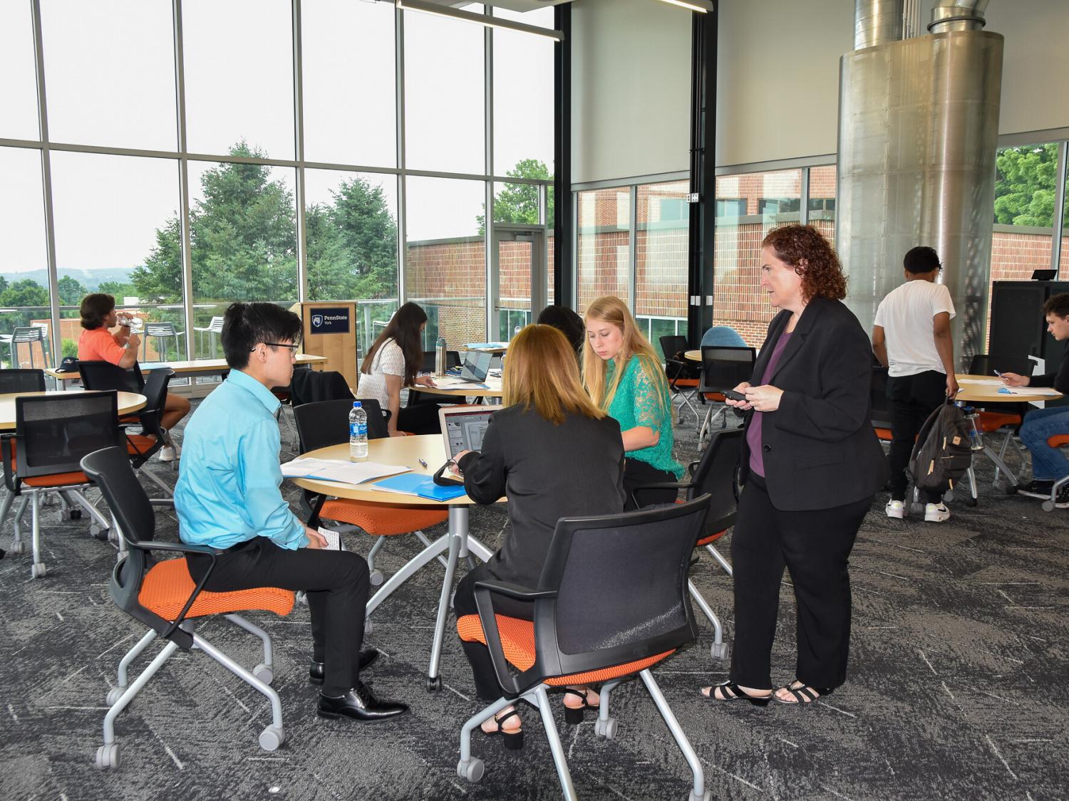 Faculty member  and groups of students arouind tables discussing work