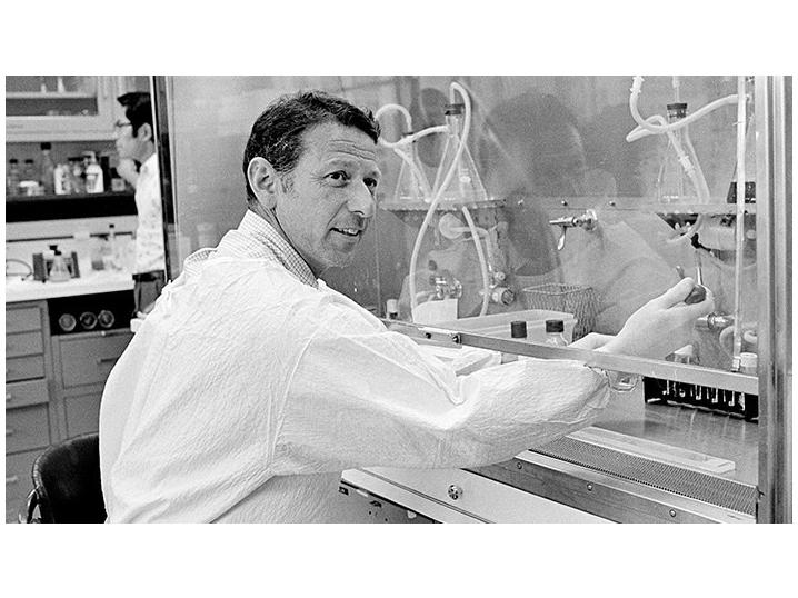 Paul Berg in the lab, working on an experiment using a lot of equipment behind glass. He's wearing a white lab coat and smiling at the camera.
