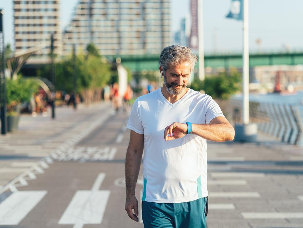 A middle-aged, bearded man wearing a short sleeve shirt walks along an urban waterfront while looking at his wearable device on his wrist. 