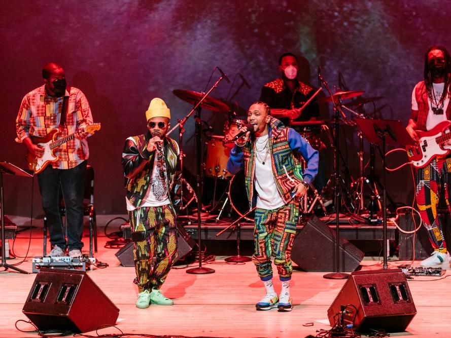 Two men of color sing into microphones while a band plays behind them.