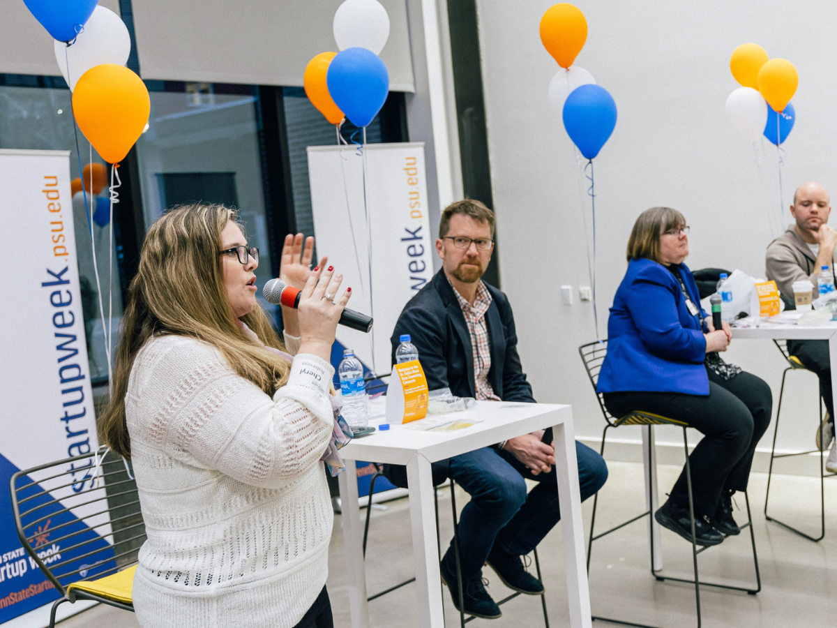 Four individuals speak at the front of a room with Startup Week branding and balloons around them.