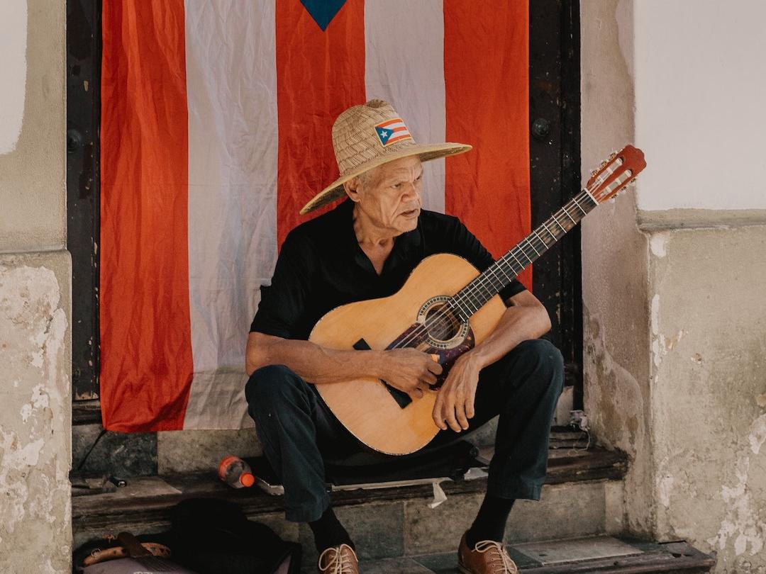 Elderly man with guitar sitting in front of Puerto Rican flag.