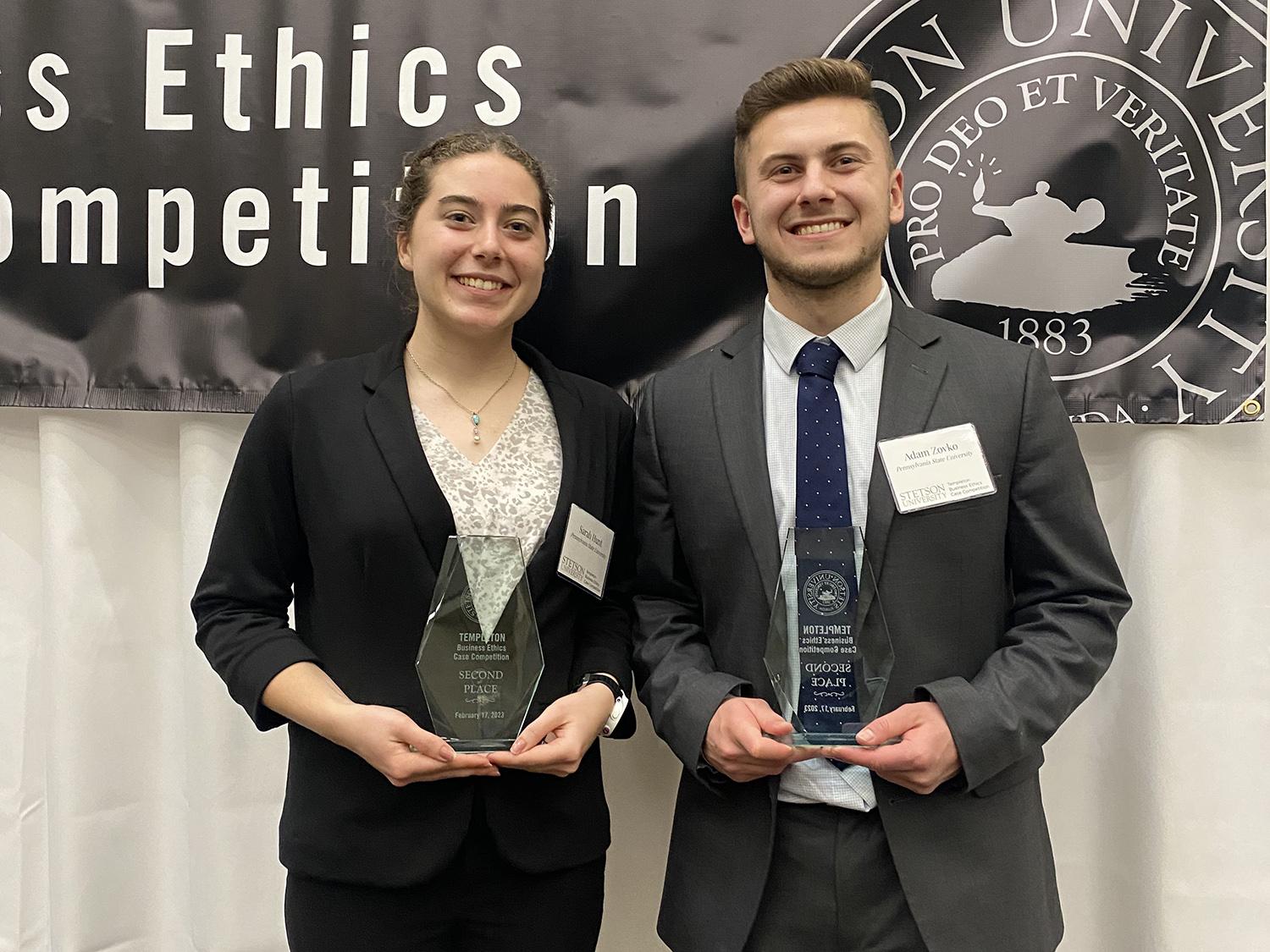 A young woman and young man in formal dress pose with trophies.