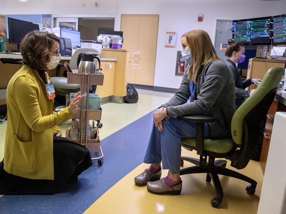 A woman in a cardigan sweater kneels in a hospital common room. She speaks with a woman in scrubs and a sweater sitting in a chair in front of her. Next to the kneeling woman is a rack of coffee pots and other implements. Behind the seated woman in a bank of computer screens, and another woman sits at a desk. Everyone is masked.