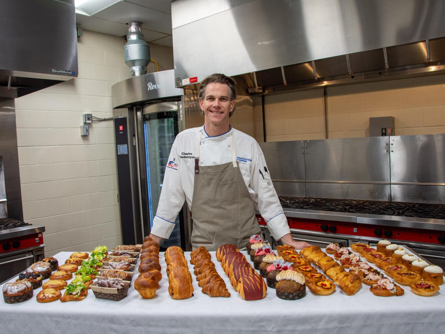 Chef Charles R. Niedermyer standing in a kitchen behind a table filled with rows of pastries