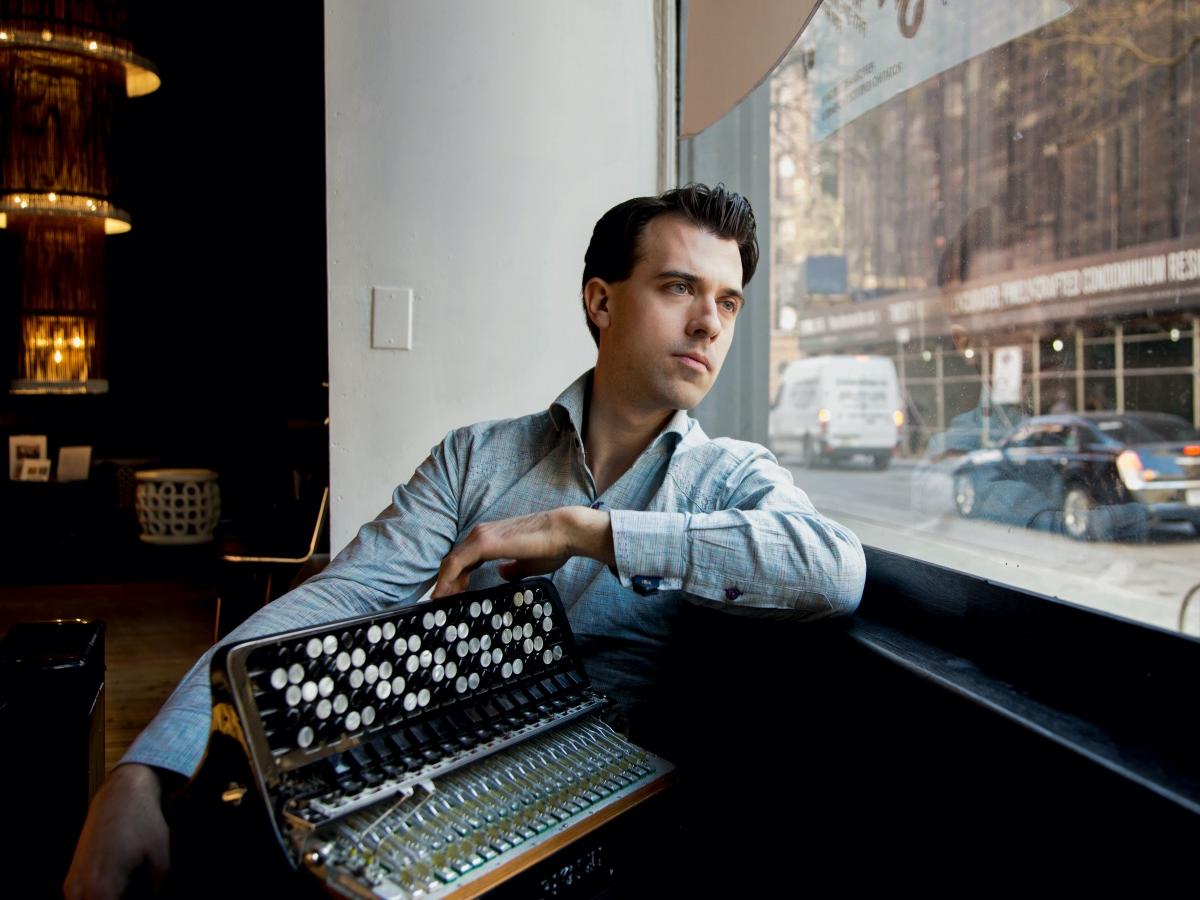 A man holding a bandoneon buttoned reed instrument sits at a window along a busy street.