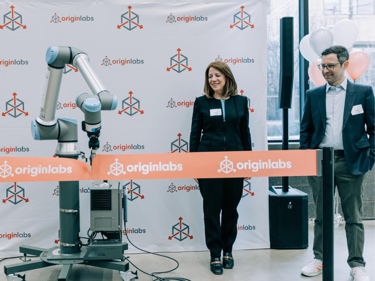 Lora Weiss and Ryan Mandell stand next to a robotic arm which is cutting the OriginLabs ribbon in front of an OriginLabs backdrop