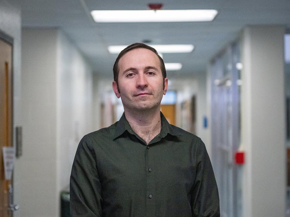 Portrait of a person in a dark green button-up shirt standing in a hallway.
