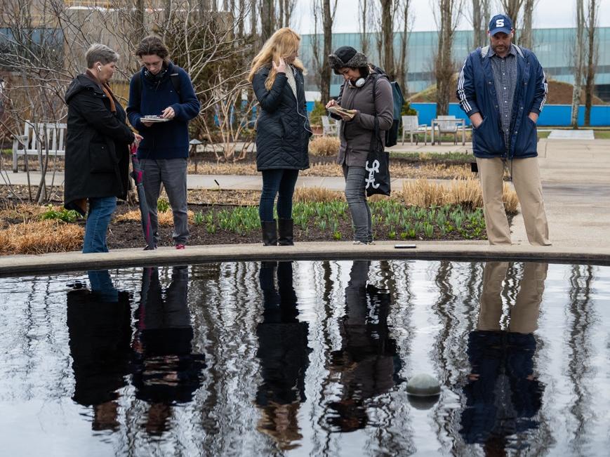 A group of five people pause in front of a reflecting pool as they listen to a prompt through headphones.