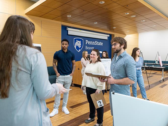 Students and library staff members discuss a student research project. The back wall is blue and displays the Penn State Beaver logo in white.