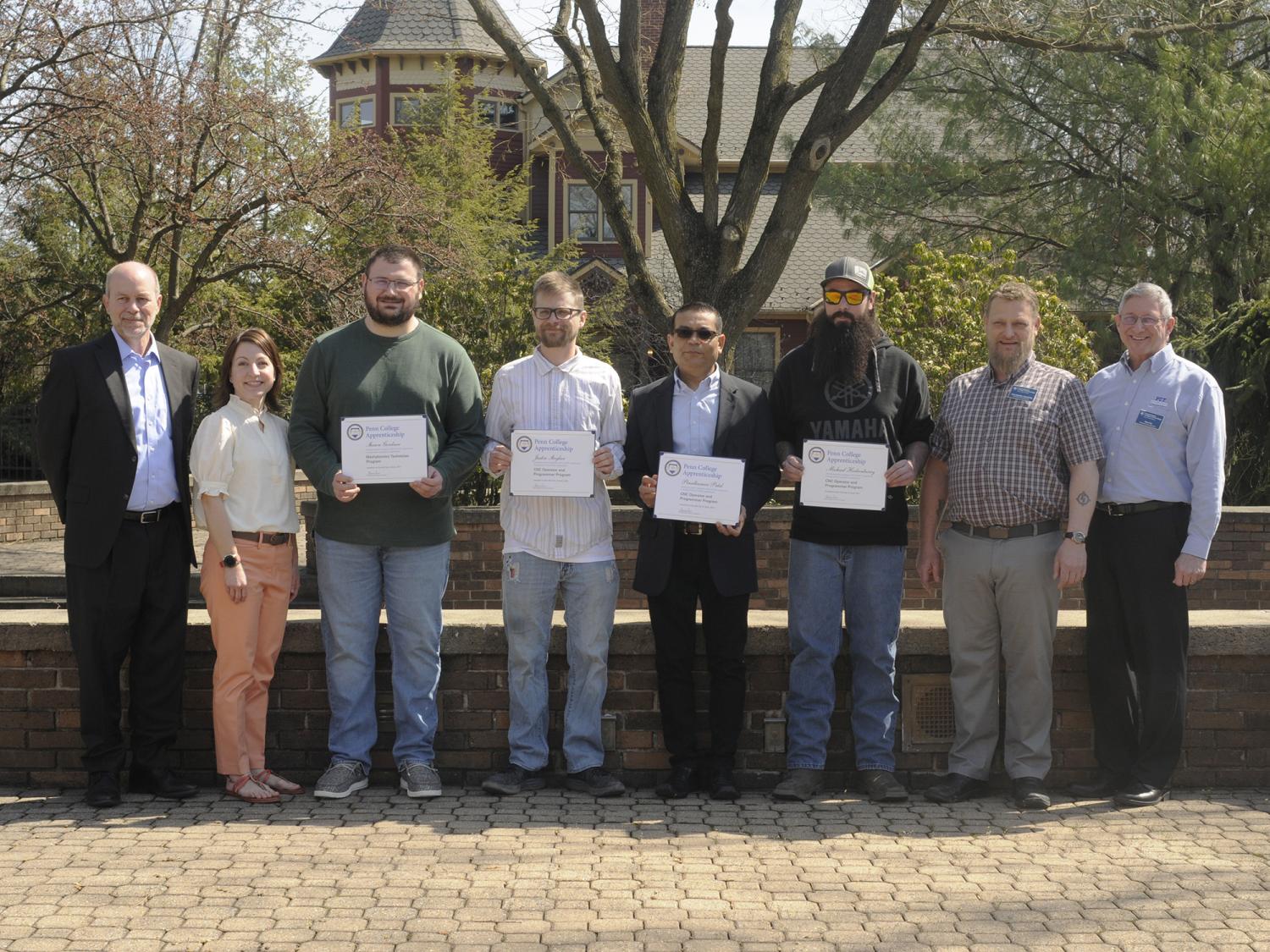 A group of eight people stand in a line near The Victorian House on the Penn College campus. Some are holding certificates.