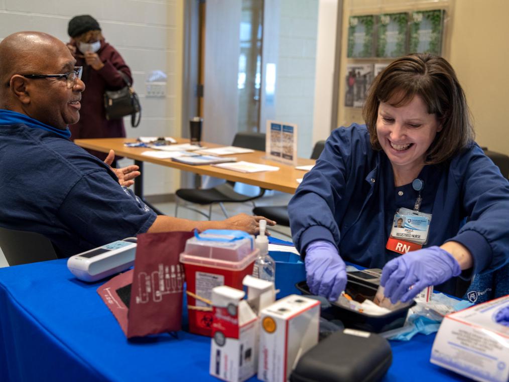 A man and woman sit at a table. The woman, a nurse, is providing a health screening. 
