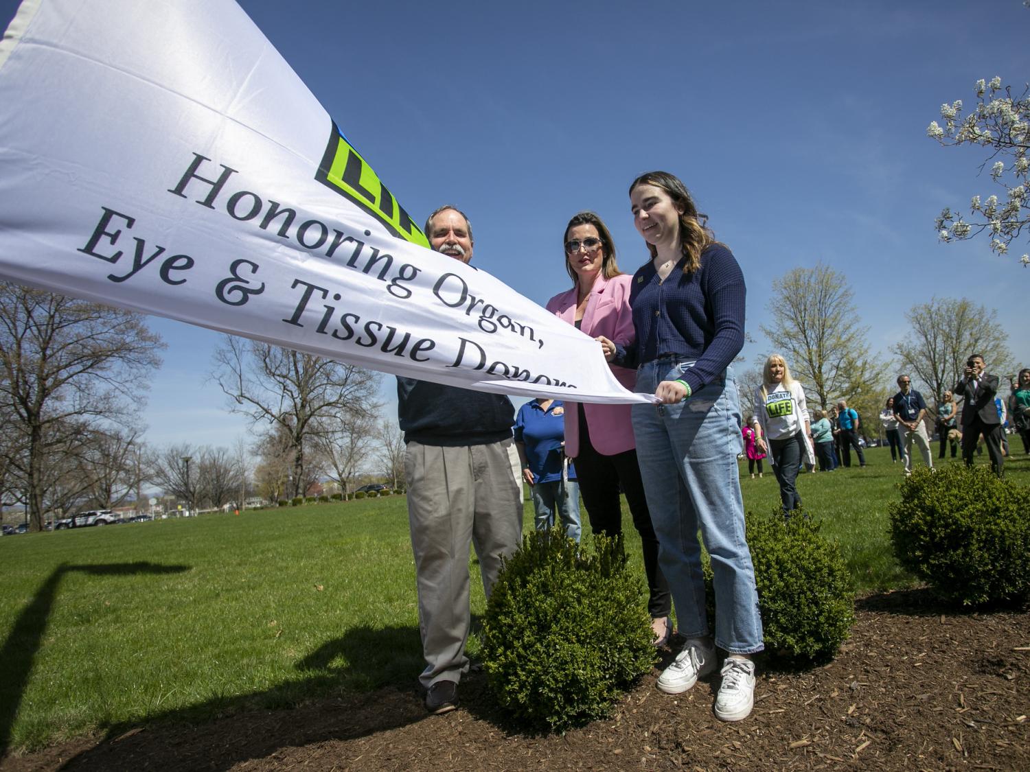 A man, a woman and a teenage girl hold a “Donate Life” flag while bystanders look on. Bushes and grass are in the foreground.