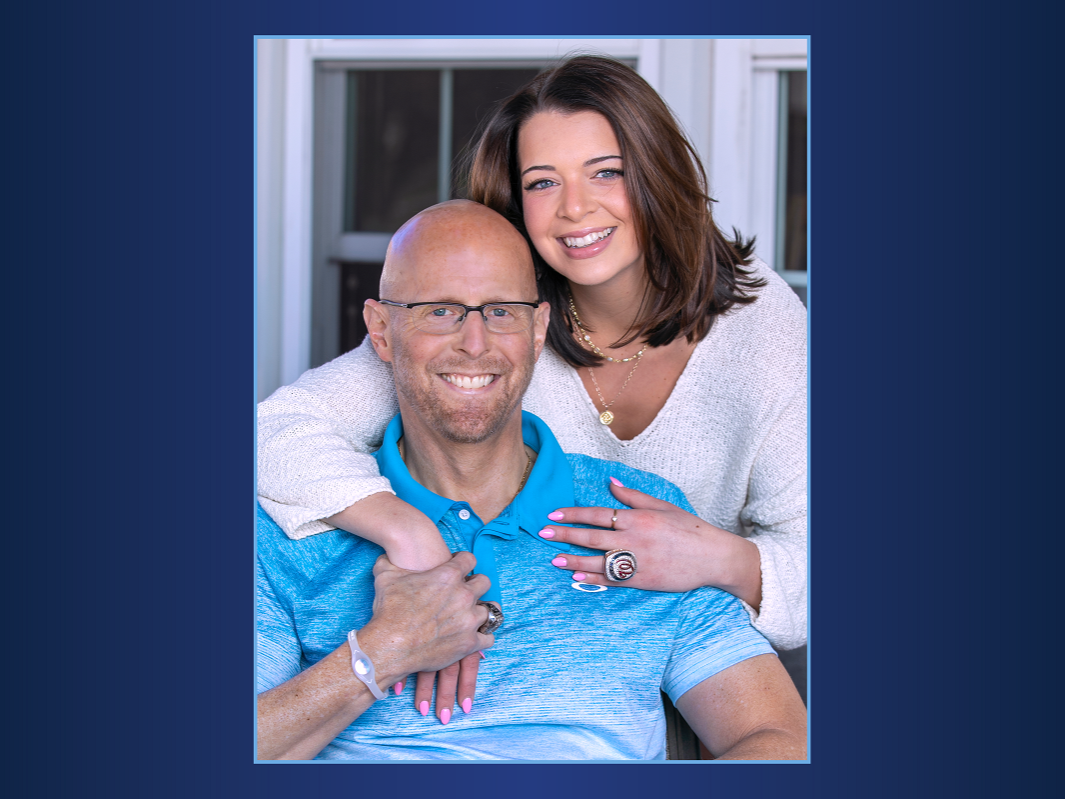 Sydney Harris, who has shoulder-length hair and wears her dad’s World Series Championship ring on her left hand, smiles as she encircles him in a hug. Doug, who is bald, is seated in front of Sydney and has a short beard, glasses and big smile on his face.
