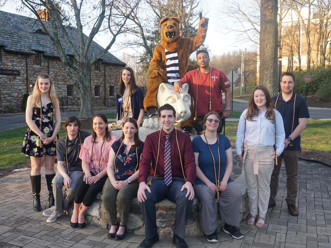 Group of students seated surrounding statue of Nittany Lion in grass field.