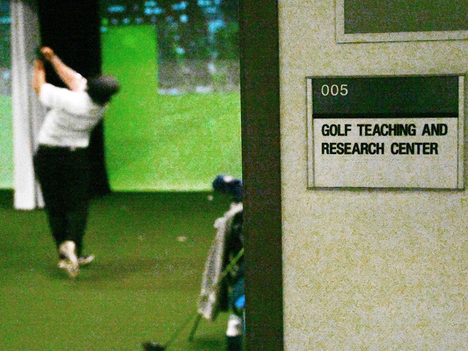 Looking from outside room through doorway with sign visible on right, a golfer inside the room hits a ball into a screen.