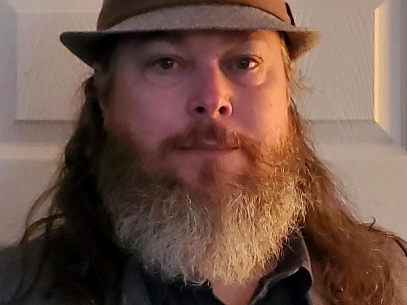 Man with a grey beard wearing tan fedora-style hat.